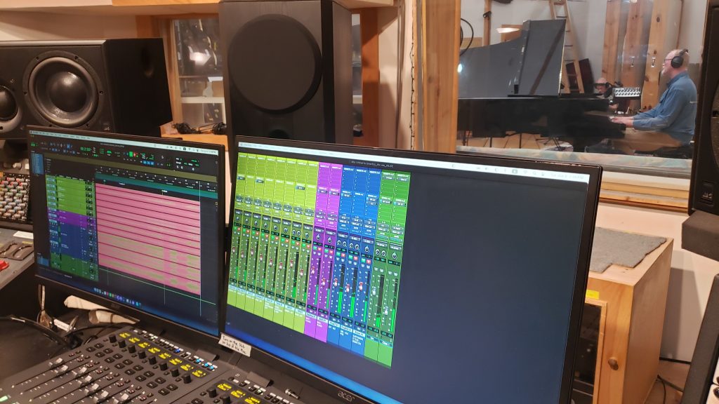 Two computer monitors showing Pro Tools and Eric Lindahl at piano through window.